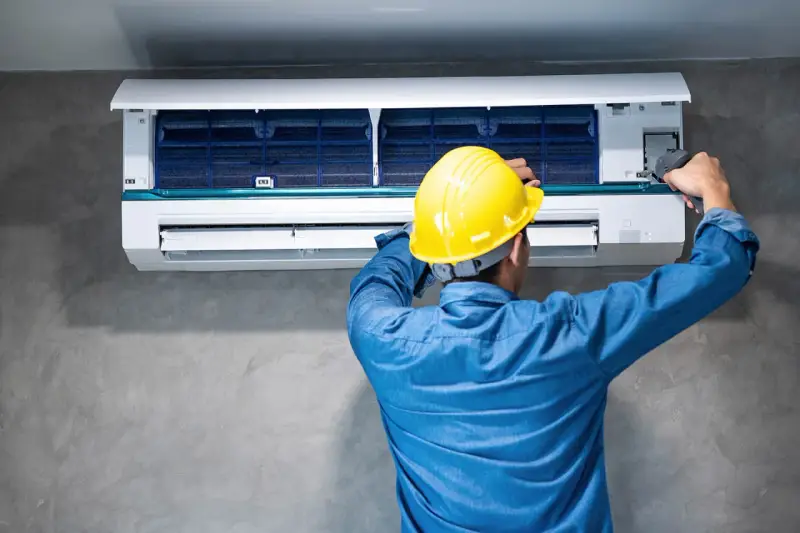 Técnico con casco amarillo revisa la unidad interior de aire acondicionado, buscando la causa del ruido.
